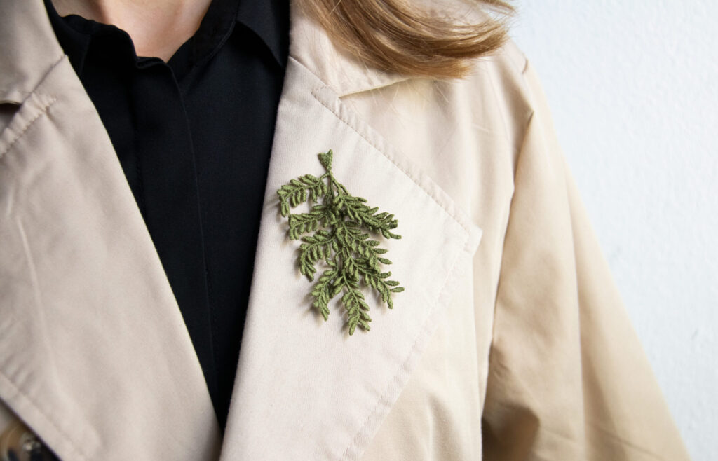 Olive green machine embroidered brooch in a shape of an olive tree branch is hanging from a collar of a beige coat